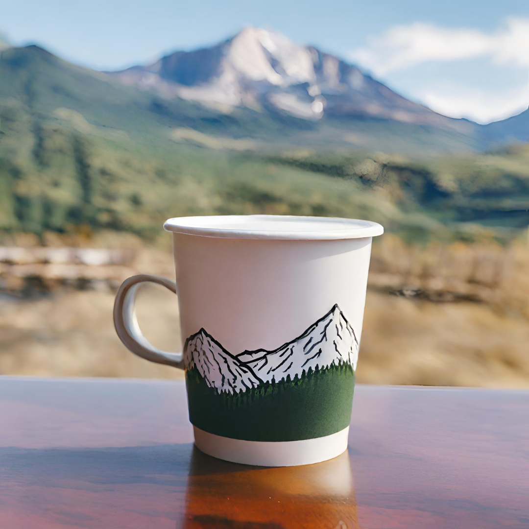 White enamel coffee mug with a mountain illustration sitting on a wooden table, with a scenic mountain landscape blurred in the background, representing outdoor coffee moments from Rock Creek Coffee Roasters.