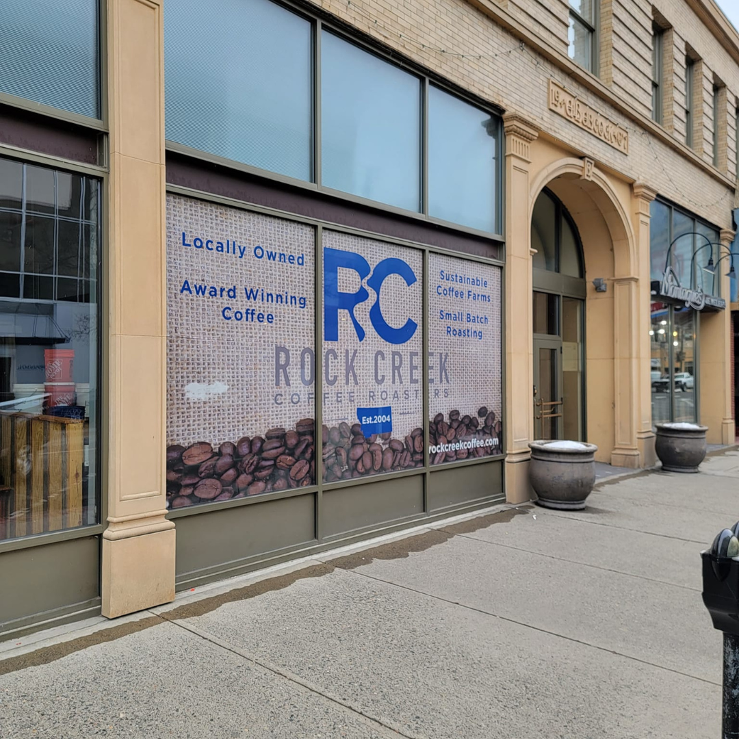 Exterior storefront of Rock Creek Coffee Roasters in downtown Billings, Montana, featuring large window signage with the RC logo, locally owned and award-winning coffee messaging, and a sidewalk view of the historic building facade.