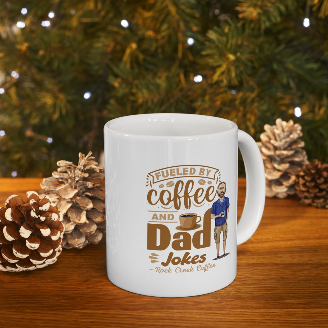 White Rock Creek Coffee mug with “Fueled by Coffee and Dad Jokes” design displayed on a wooden table with pinecones and a softly lit Christmas tree in the background.