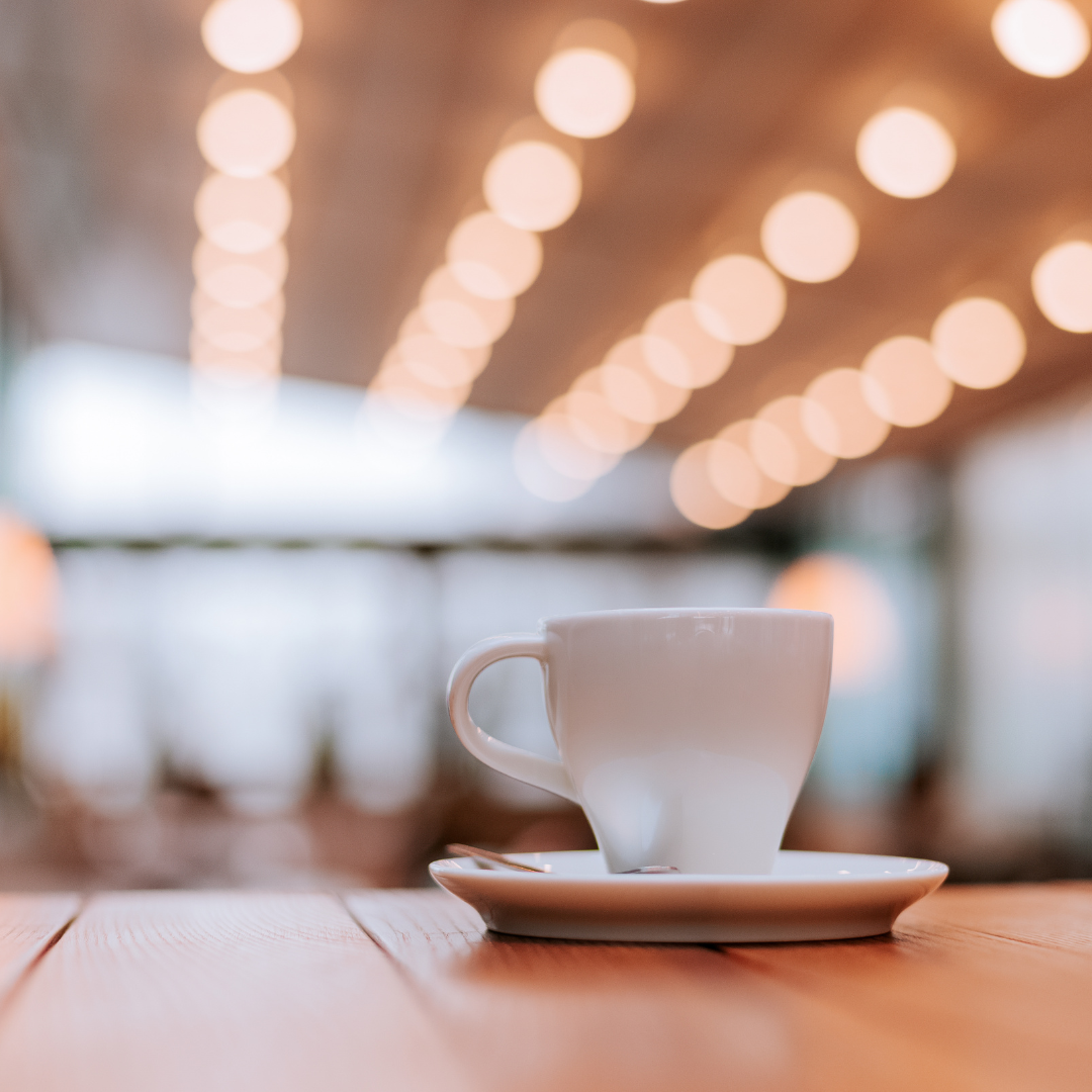 White coffee cup on a saucer placed on a wooden table with warm, blurred café lights in the background.