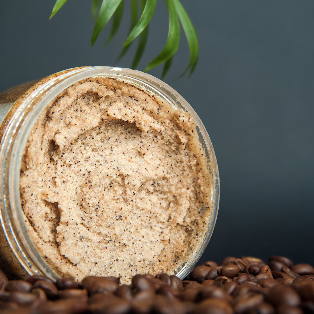 Close-up of an open jar of coffee scrub resting on roasted coffee beans with a green leaf above