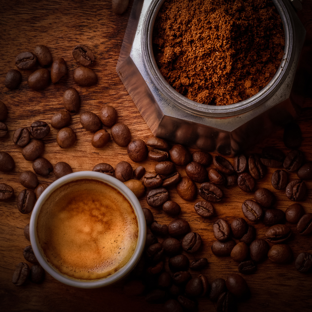 Top-down view of an espresso cup with crema surrounded by whole coffee beans and an open moka pot filled with ground coffee on a wooden table