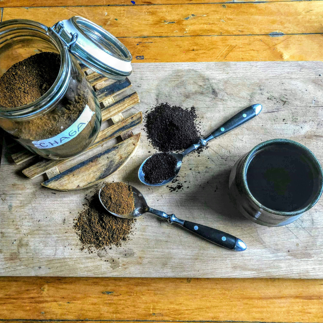 Open jar of chaga powder with two spoons of ground coffee and a cup of dark coffee on a wooden board.