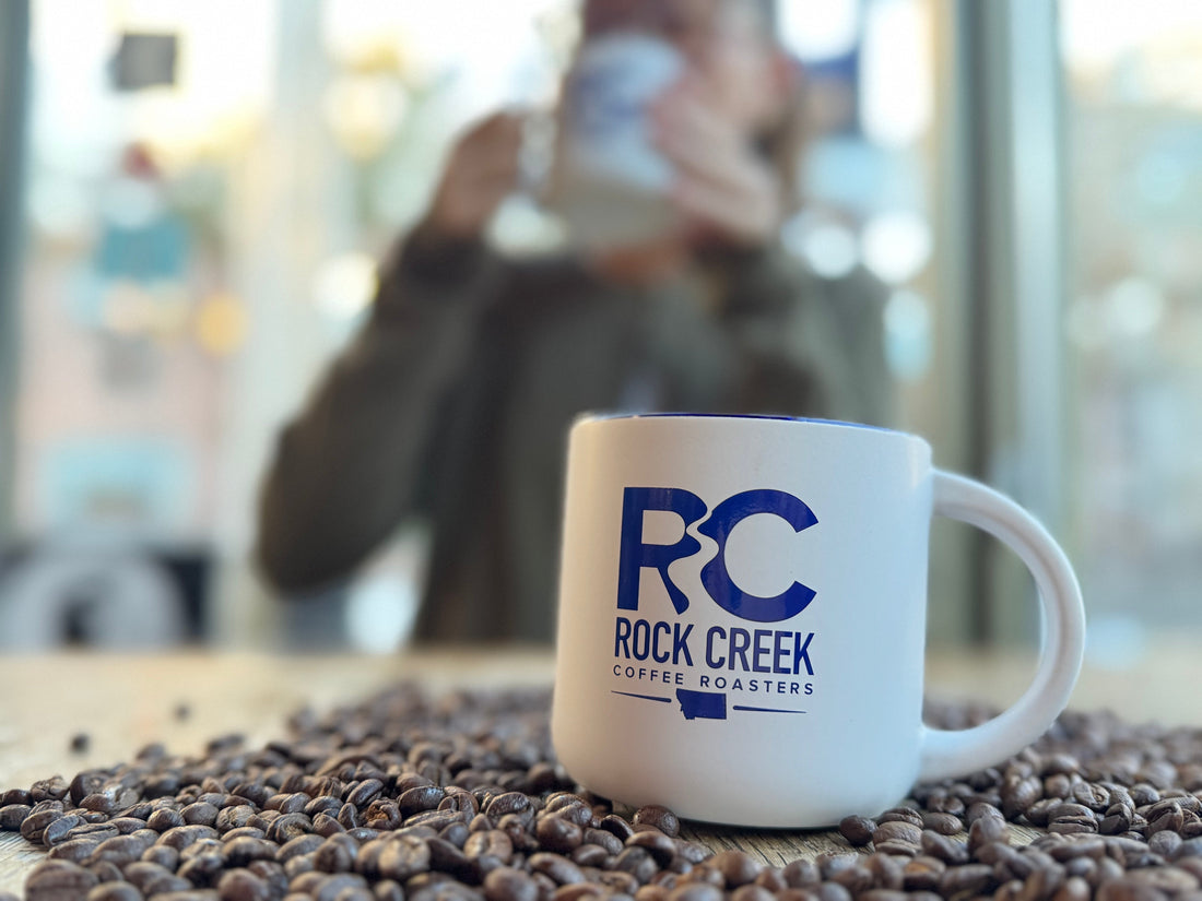 White Rock Creek Coffee Roasters mug on a table covered with roasted coffee beans, with a person blurred in the background holding a mug inside a bright café setting.