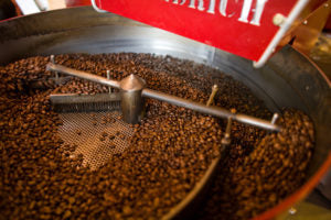 Coffee beans roasting inside a commercial drum roaster, with a metal stirring arm rotating over freshly roasted beans at Rock Creek Coffee Roasters.