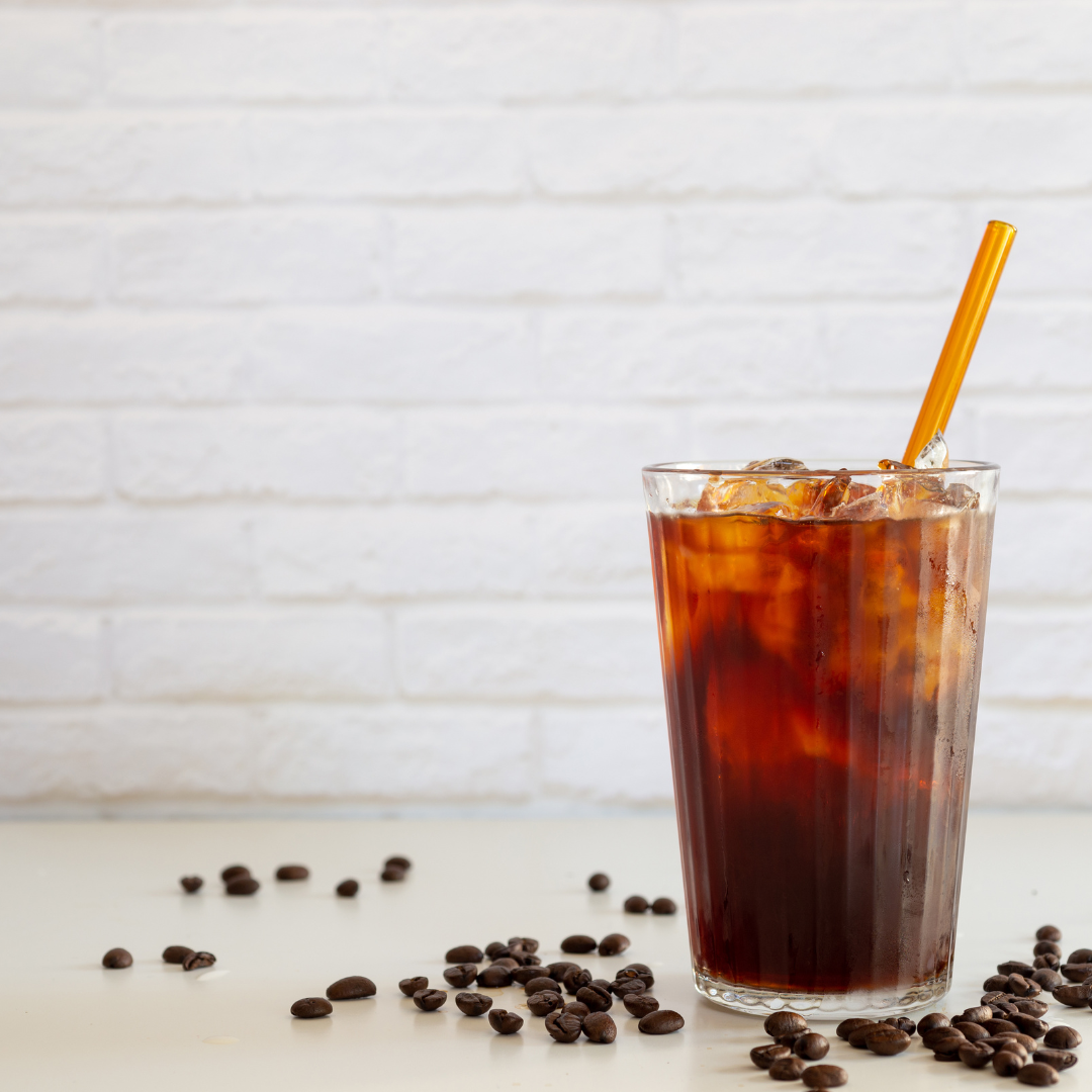 Glass of iced coffee with ice cubes and a straw, surrounded by coffee beans on a light surface