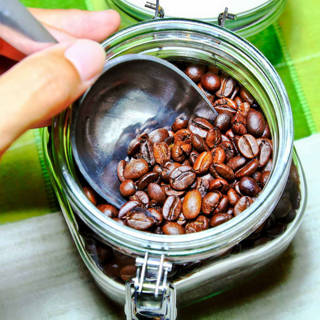 Hand holding a metal scoop inside a glass jar filled with roasted coffee beans on a green cloth