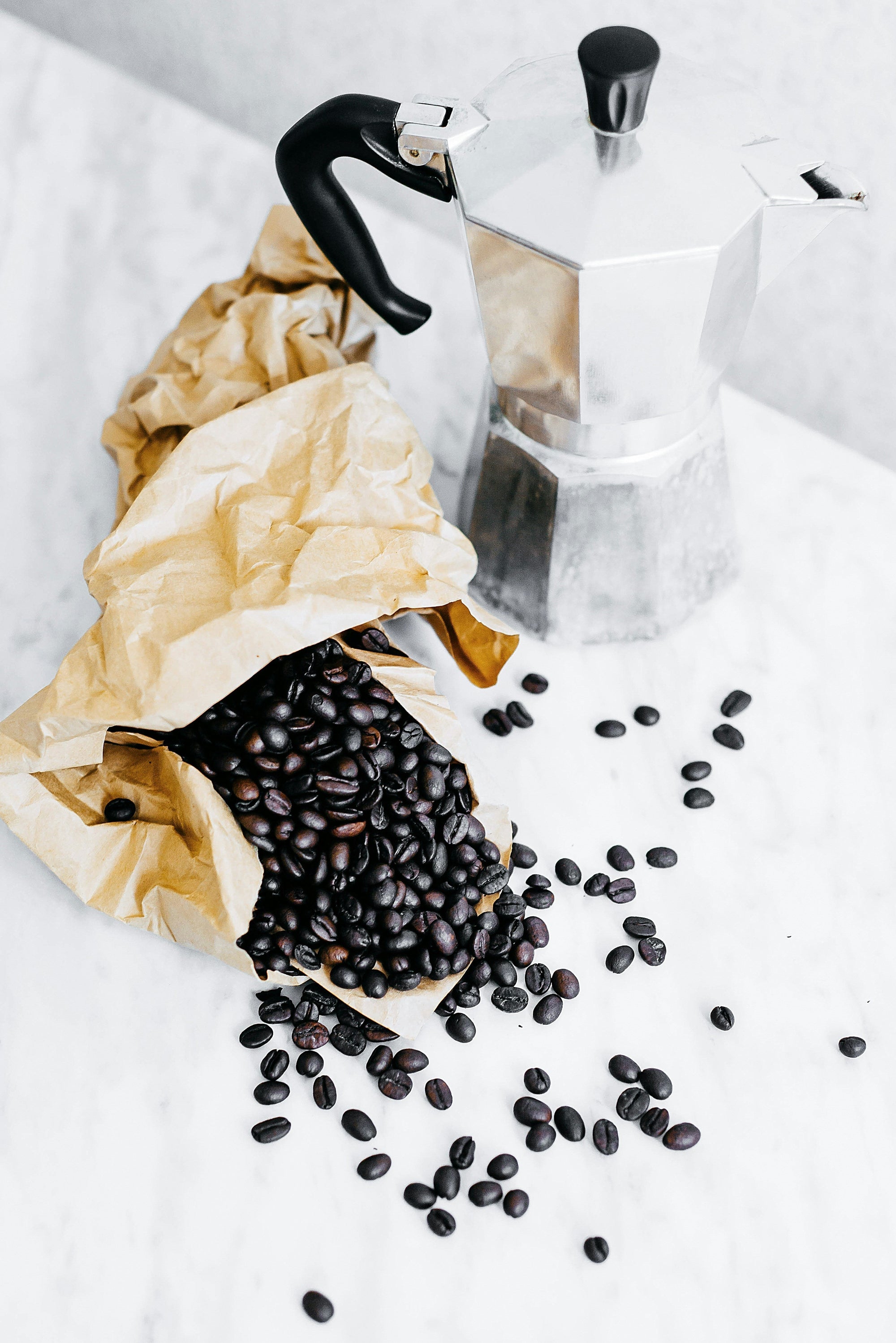 Aluminum stovetop moka pot beside a crumpled paper bag spilling roasted coffee beans onto a light marble countertop