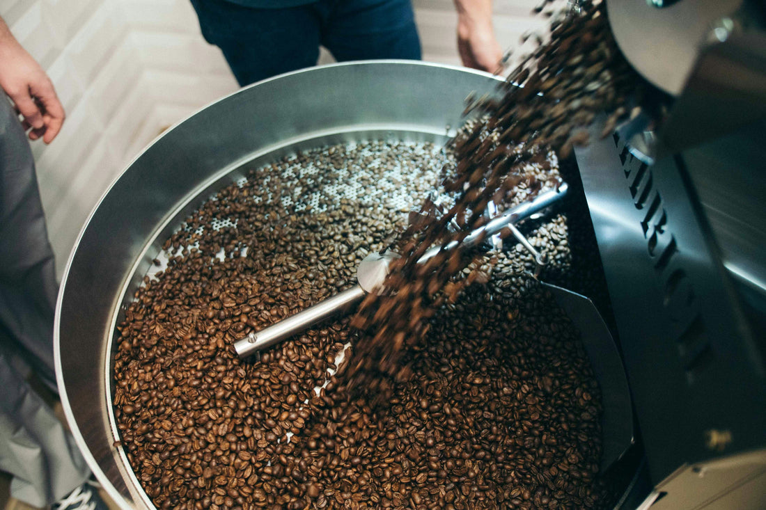 Freshly roasted coffee beans cascading into a large stainless steel cooling tray inside a commercial roaster, with beans tumbling and stirring as part of the roasting process