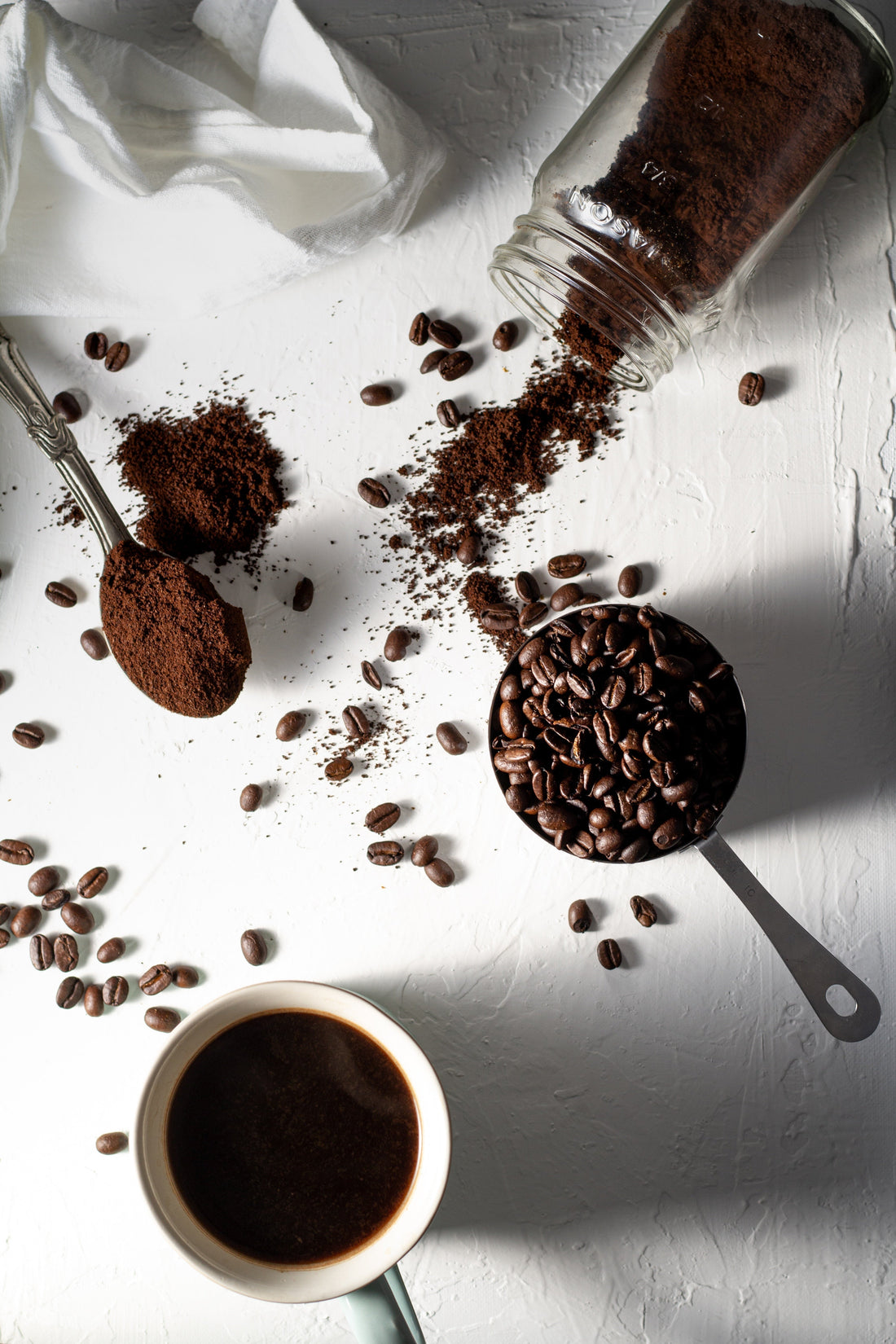 Flat lay of ground coffee spilling from a glass jar onto a white surface, with whole coffee beans scattered around, a metal scoop filled with beans, a spoonful of grounds, and a cup of black coffee nearby.