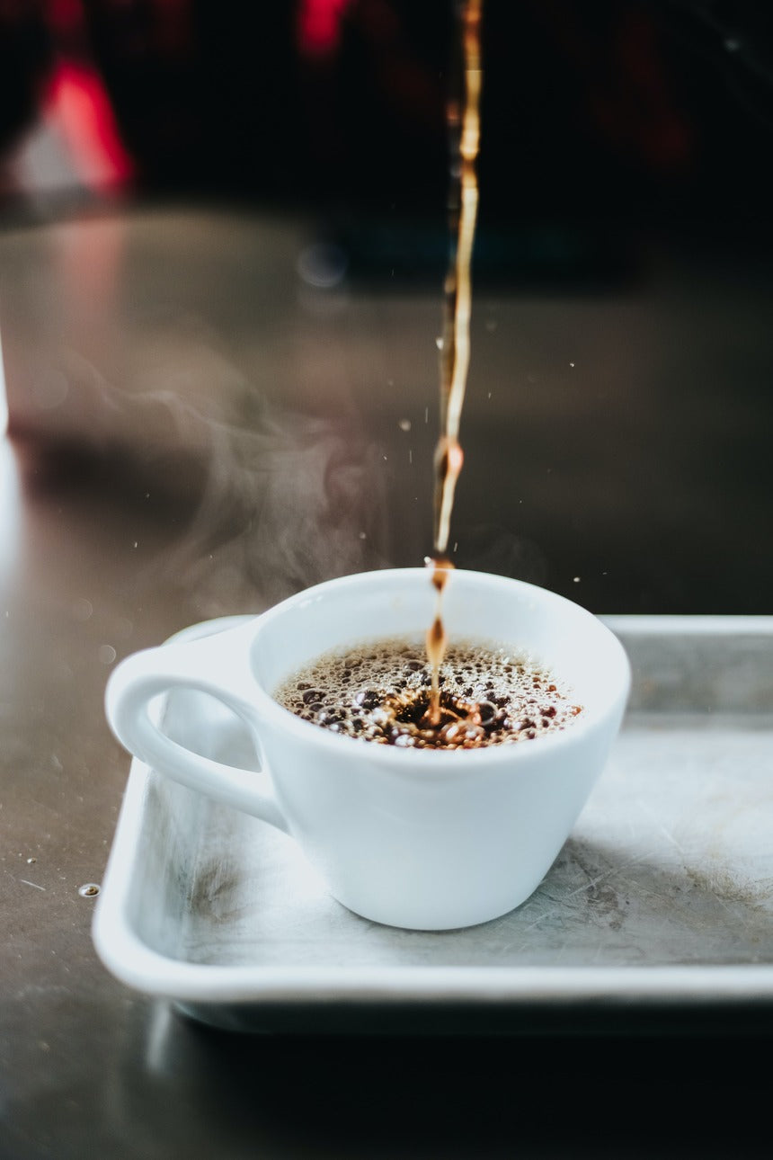 Freshly brewed hot coffee being poured into a white ceramic cup on a metal tray, with steam rising to highlight aroma and warmth in a coffee shop setting.