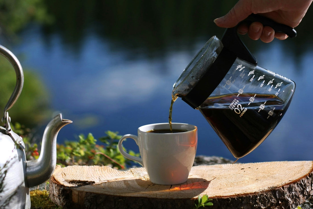 Hand pouring freshly brewed coffee from a glass carafe into a white mug on a tree stump beside a metal kettle with a calm blue lake in the background, capturing a peaceful outdoor coffee moment in nature