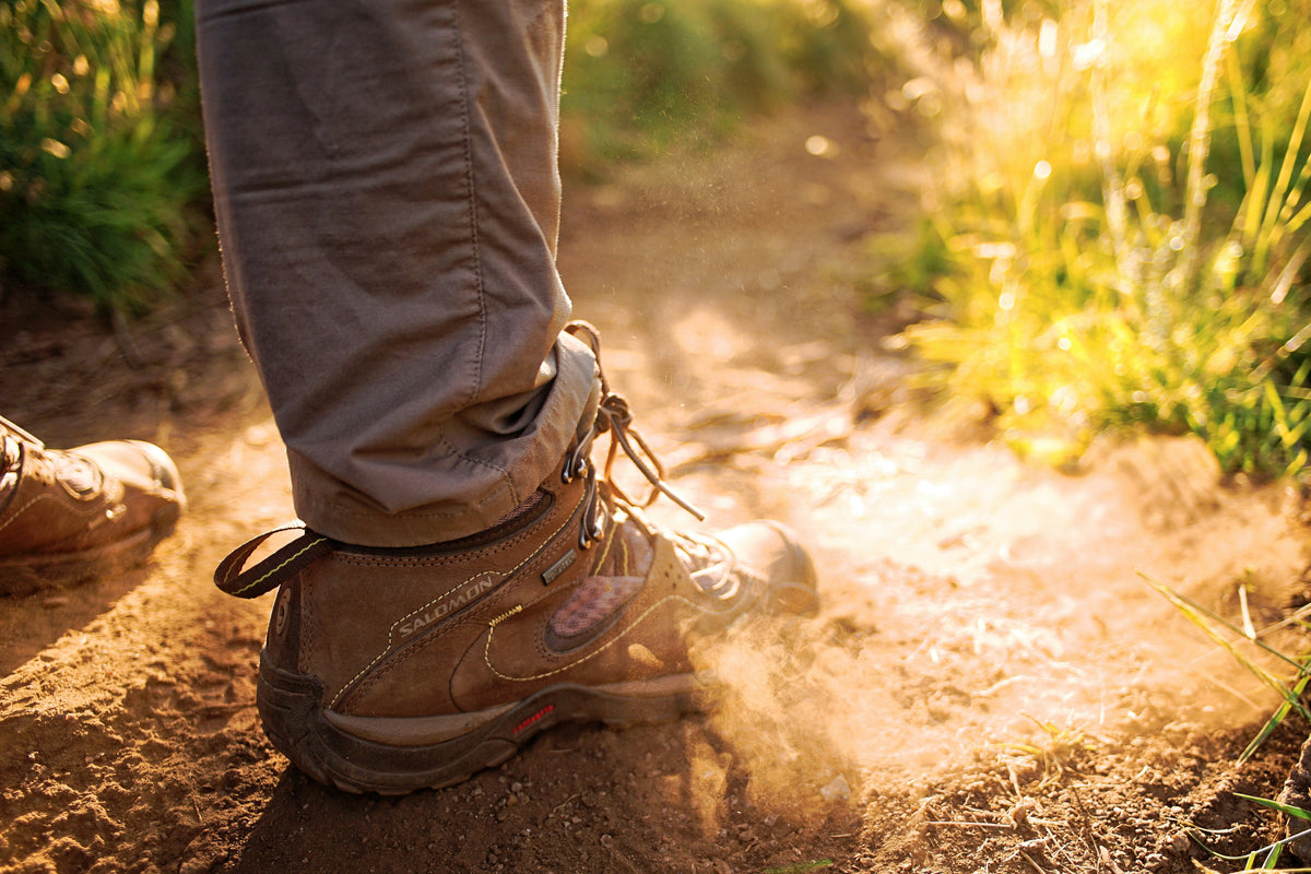 Close-up of hiking boots kicking up dust on a sunlit trail surrounded by grass