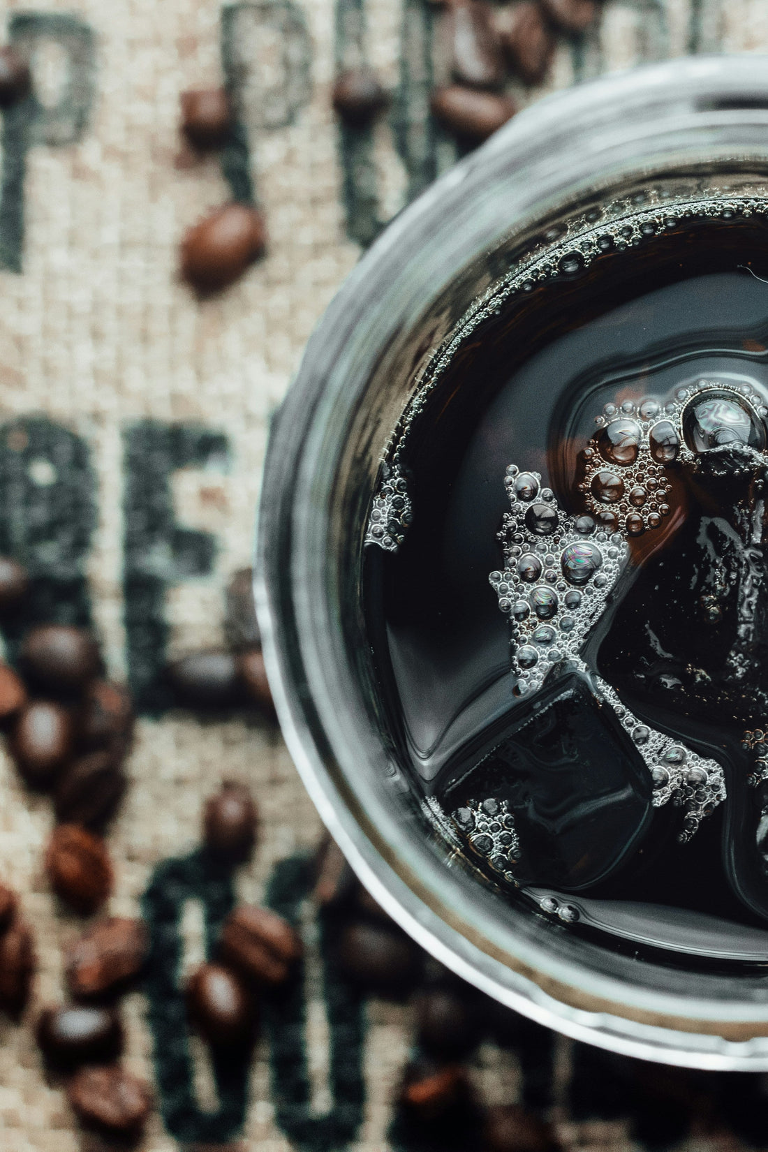 Top-down close-up of a glass of iced black coffee with visible ice cubes and bubbles, set on burlap fabric scattered with whole coffee beans