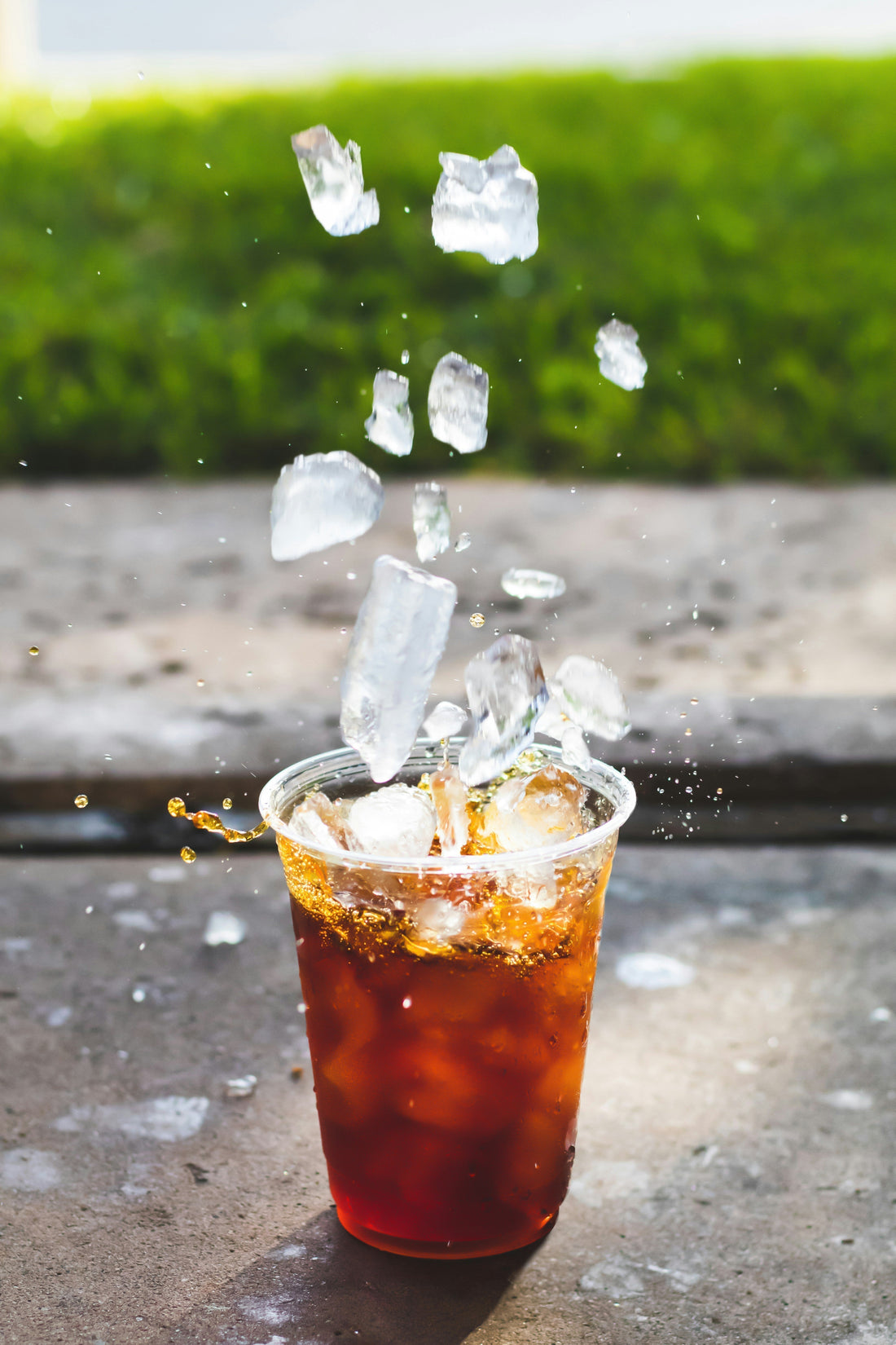 Iced coffee in a clear plastic cup with ice cubes splashing into the drink outdoors