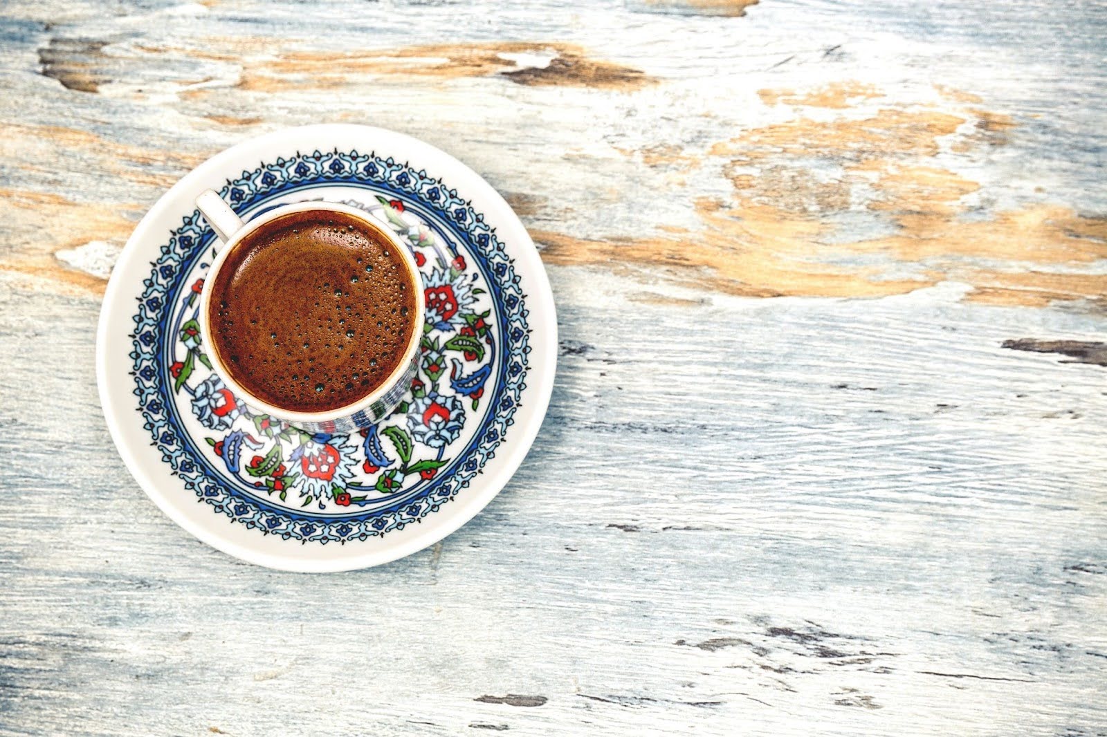 Overhead view of a freshly brewed cup of coffee on a colorful patterned saucer, placed on a rustic whitewashed wooden surface