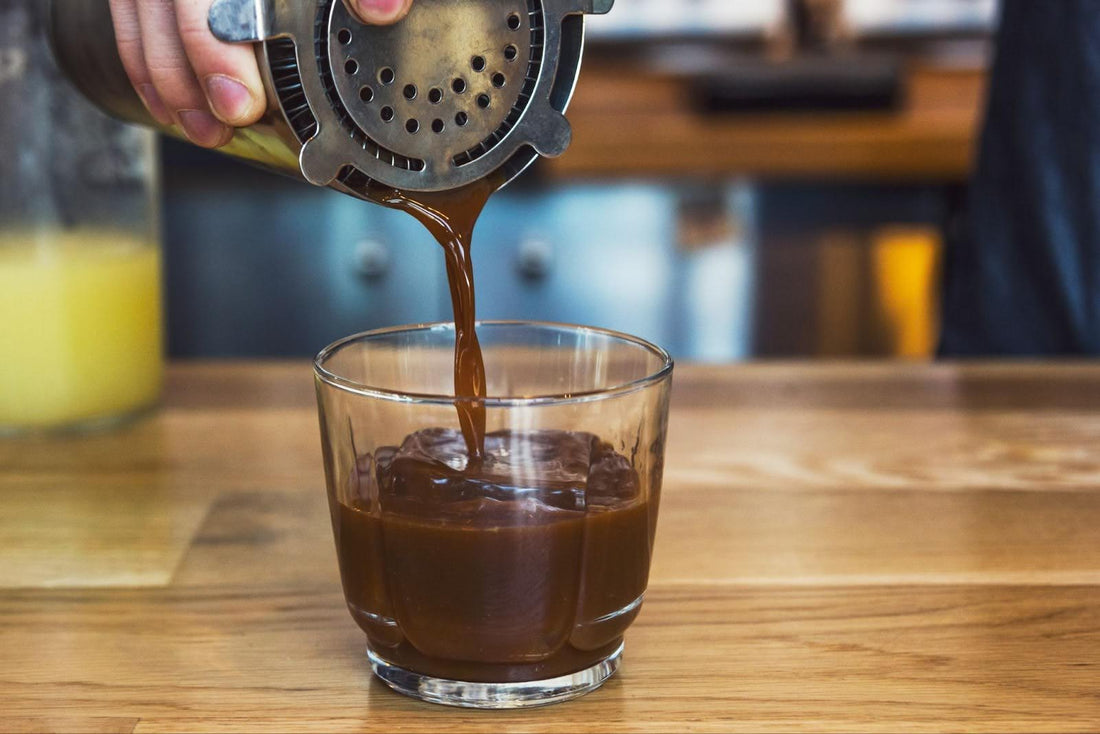 Hot coffee streaming from a French press into a clear glass filled with ice cubes on a wooden countertop, with a softly blurred café background.