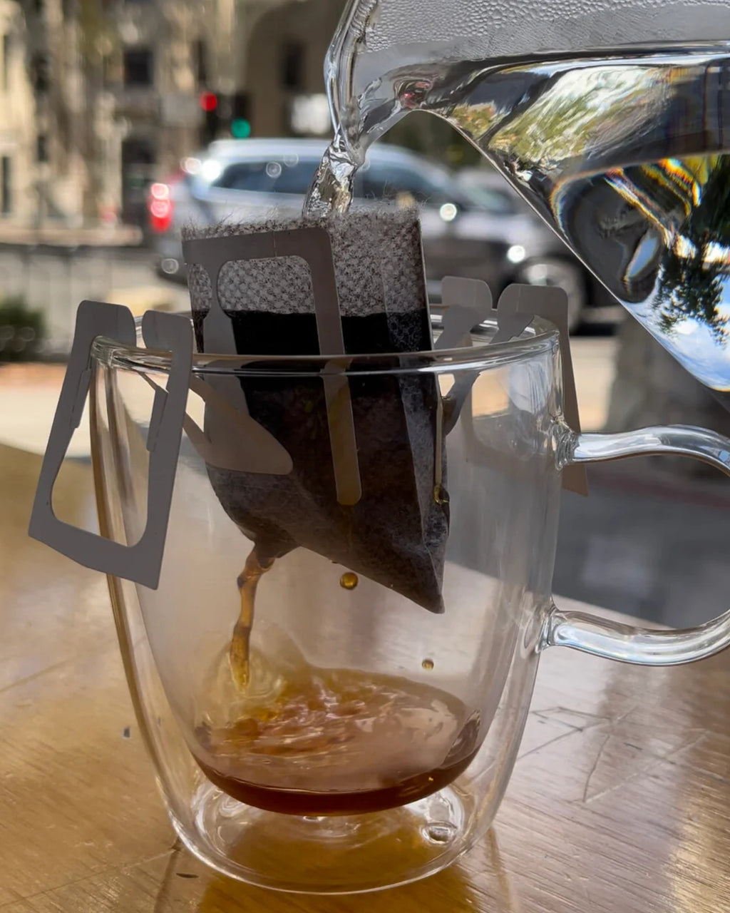 Hot water being poured from a glass kettle into a single-serve pour-over coffee filter hanging over a clear double-walled glass mug, with brewed coffee dripping into the cup at an outdoor café setting.