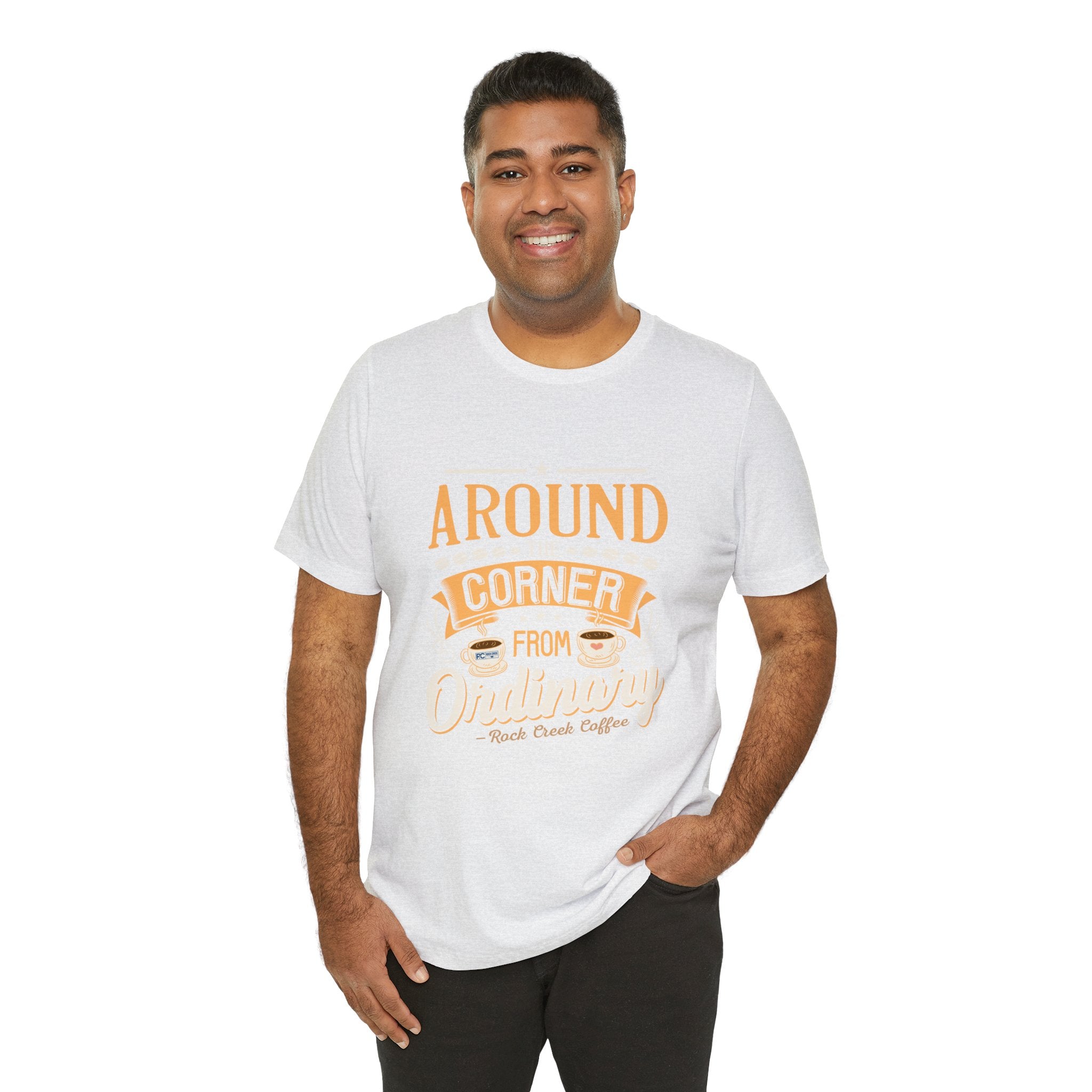 Adult man smiling while modeling an ash short-sleeve T-shirt featuring the “Around the Corner from Ordinary” graphic in a coffee-themed typographic design, shown against a plain white background.
