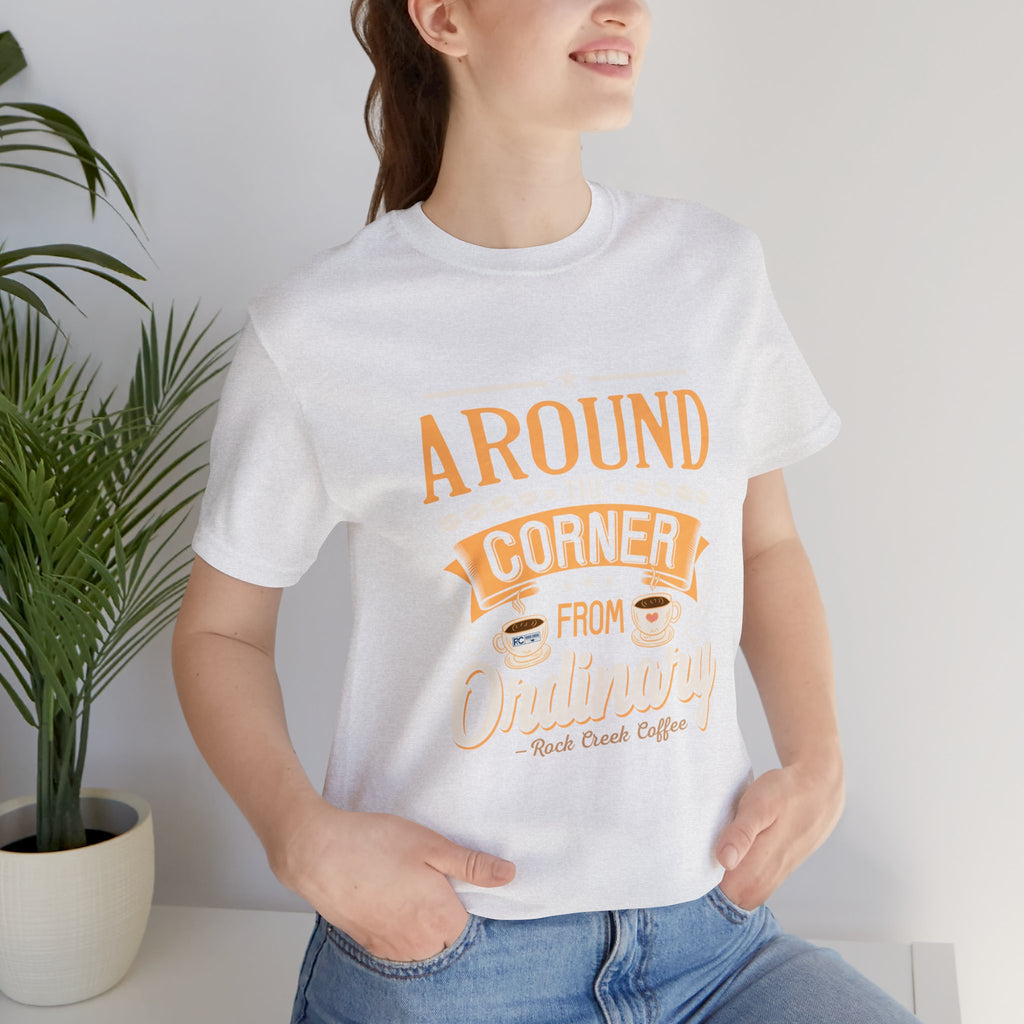 Woman wearing an ash Rock Creek Coffee T-shirt with the “Around the Corner From Ordinary” graphic in a casual indoor lifestyle photo