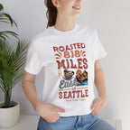 Smiling woman seated indoors modeling a white short-sleeve T-shirt printed with a coffee-themed graphic reading “Roasted 818 Miles East of Seattle,” featuring a steaming coffee cup and scenic mountain–lake illustration, with potted plants visible in the background.