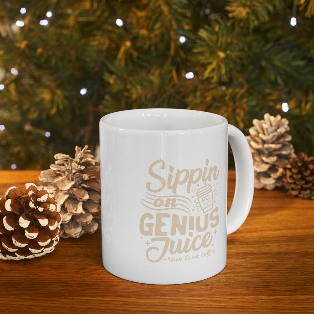 White Rock Creek Coffee mug with “Sippin on Genius Juice” in beige lettering and an RC coffee cup graphic, filled with steaming coffee and displayed on a wooden table with pinecones and warm holiday lights in the background.