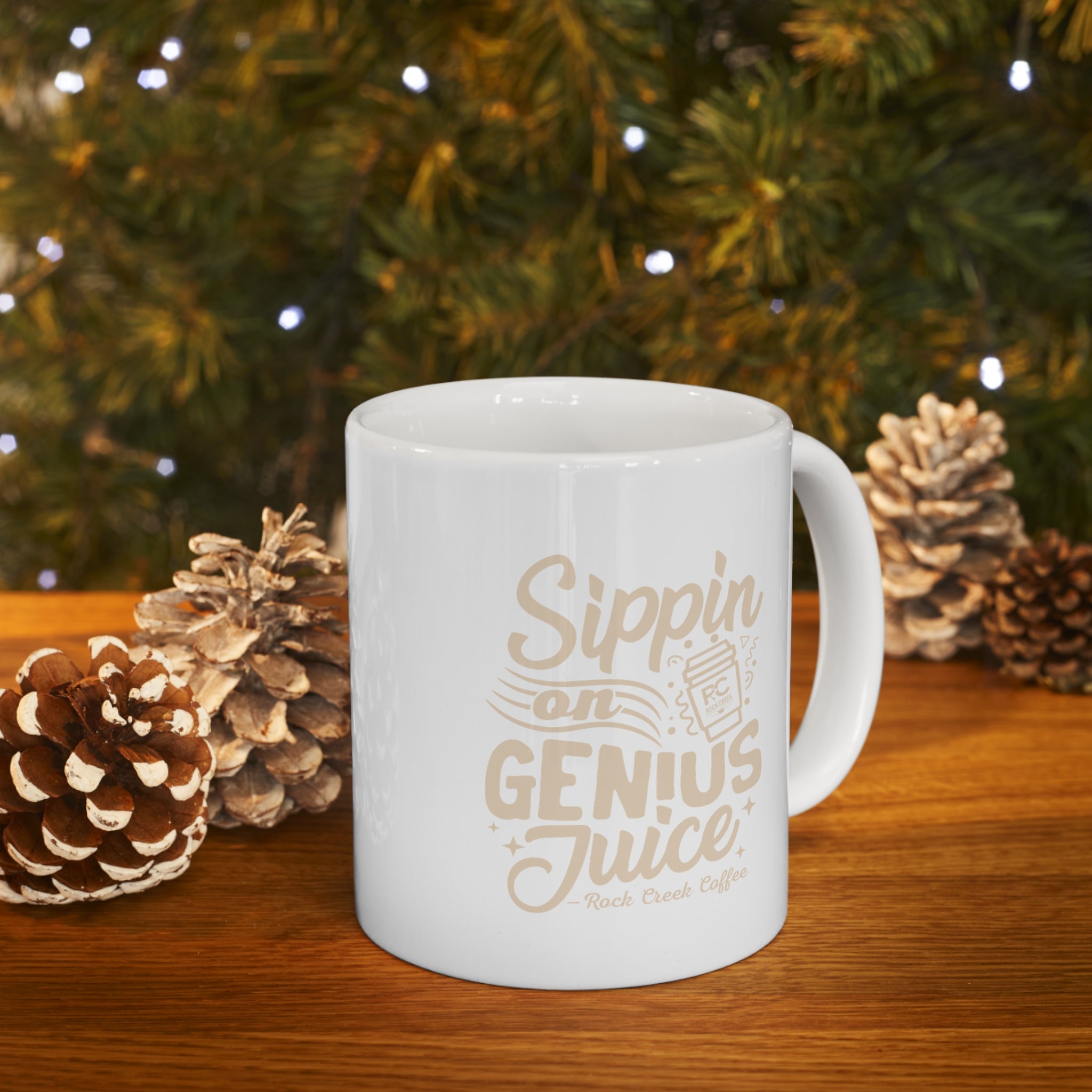 White Rock Creek Coffee mug with “Sippin on Genius Juice” in beige lettering and an RC coffee cup graphic, filled with steaming coffee and displayed on a wooden table with pinecones and warm holiday lights in the background.