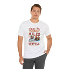 Adult man modeling a white short-sleeve T-shirt printed with a colorful coffee-themed graphic that reads “Roasted 818 Miles East of Seattle,” featuring a steaming coffee cup and mountain lake illustration, shown against a plain white background.