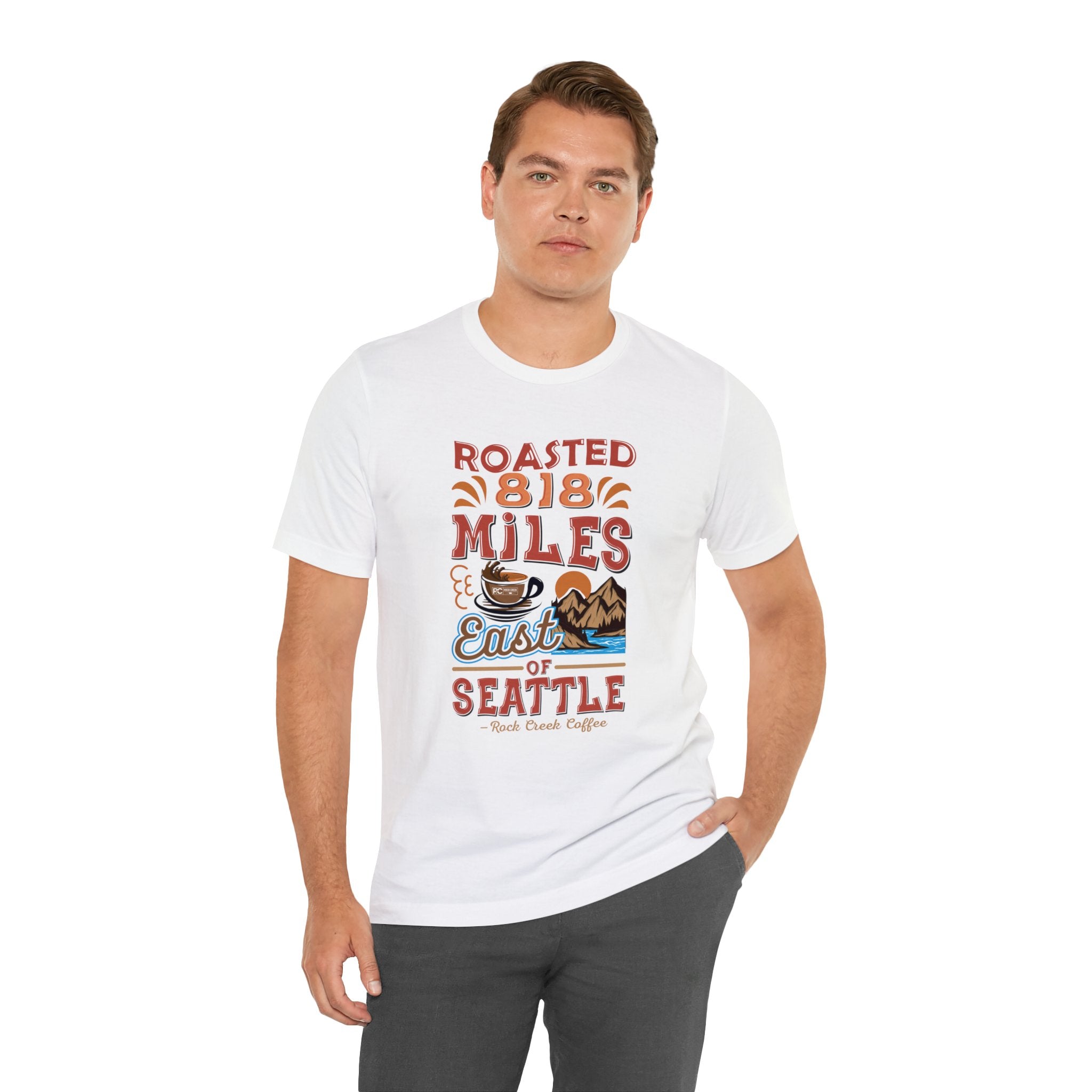 Adult man modeling a white short-sleeve T-shirt printed with a colorful coffee-themed graphic that reads “Roasted 818 Miles East of Seattle,” featuring a steaming coffee cup and mountain lake illustration, shown against a plain white background.