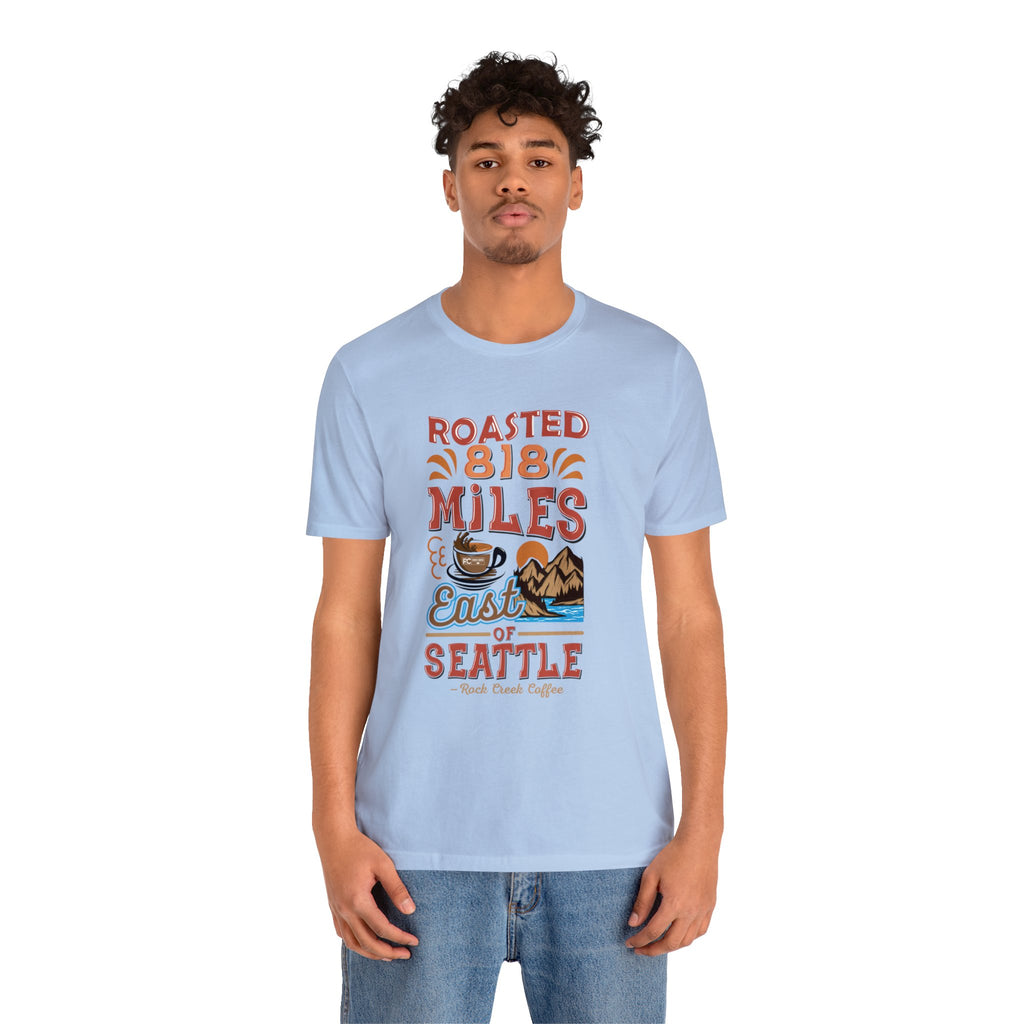 Adult man modeling a light blue short-sleeve T-shirt printed with a colorful graphic that reads “Roasted 818 Miles East of Seattle,” featuring a steaming coffee cup and mountain lake illustration, shown against a plain white background.
