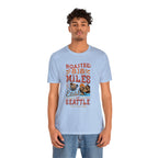 Adult man modeling a light blue short-sleeve T-shirt printed with a colorful graphic that reads “Roasted 818 Miles East of Seattle,” featuring a steaming coffee cup and mountain lake illustration, shown against a plain white background.