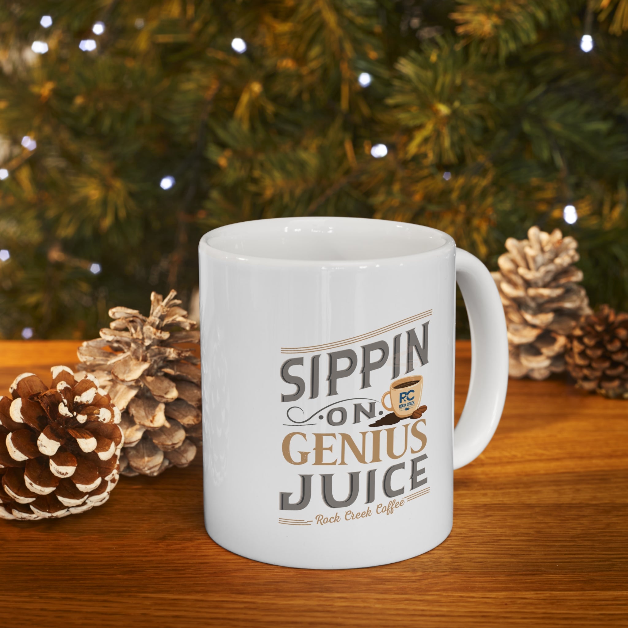 White Rock Creek Coffee mug featuring the phrase “Sippin on Genius Juice” in stylized gray and gold lettering with a small coffee cup graphic, filled with coffee and styled on a wooden surface with pinecones and warm holiday lights in the background.