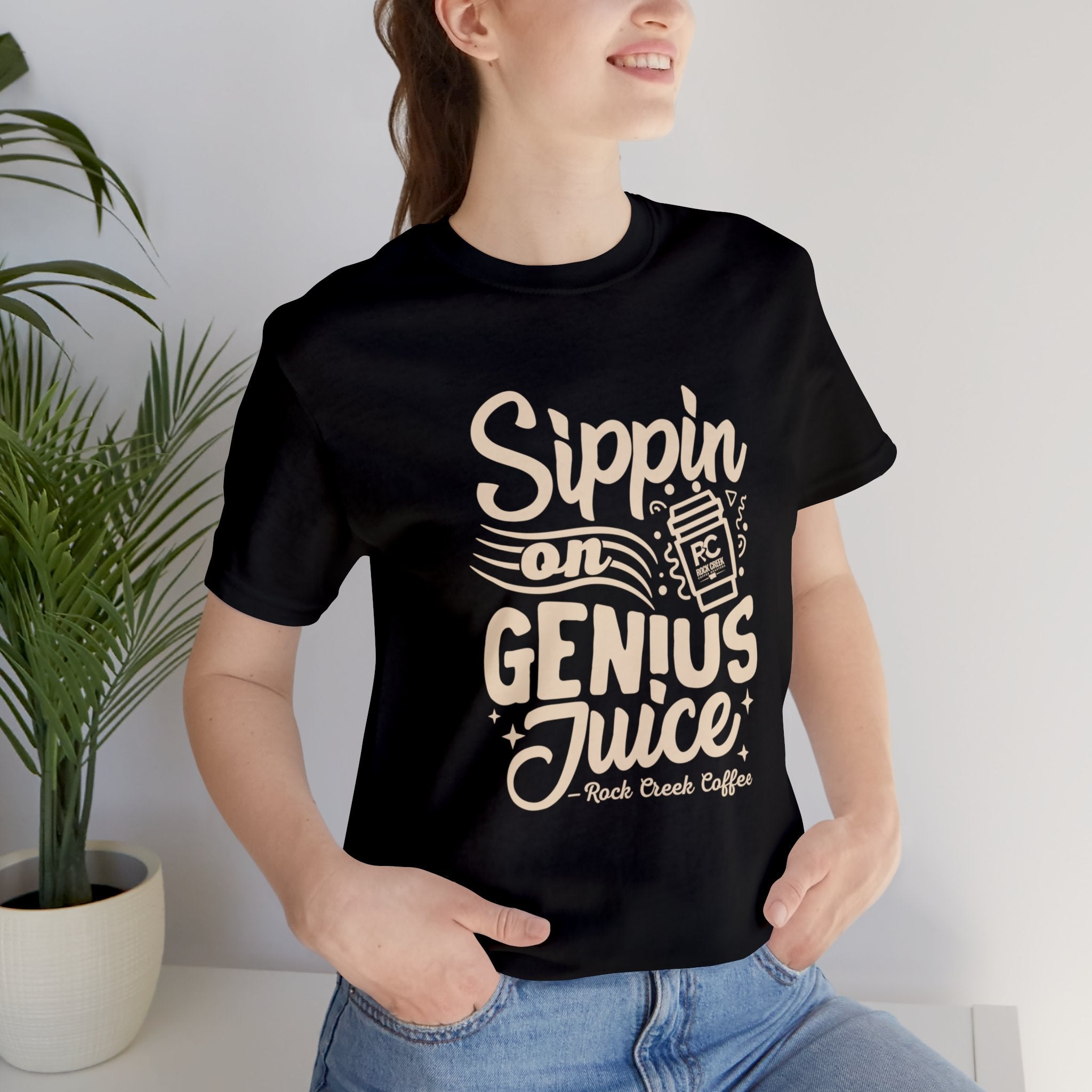 Smiling woman wearing a black short-sleeve T-shirt printed with the slogan “Sippin on Genius Juice” and a stylized to-go coffee cup graphic, seated casually indoors near a potted plant.