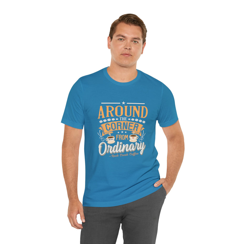 Adult man modeling an aqua short-sleeve T-shirt featuring the “Around the Corner from Ordinary” graphic in a bold coffee-themed typographic design, posed with one hand in his pocket against a plain white background.