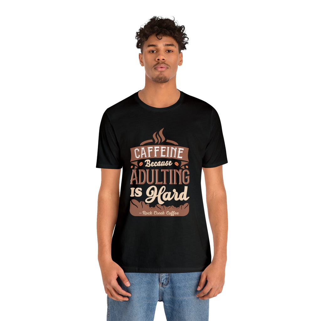 Adult man modeling a black short-sleeve T-shirt featuring the “Caffeine Because Adulting Is Hard” graphic in a stacked typographic design, shown against a plain white background.