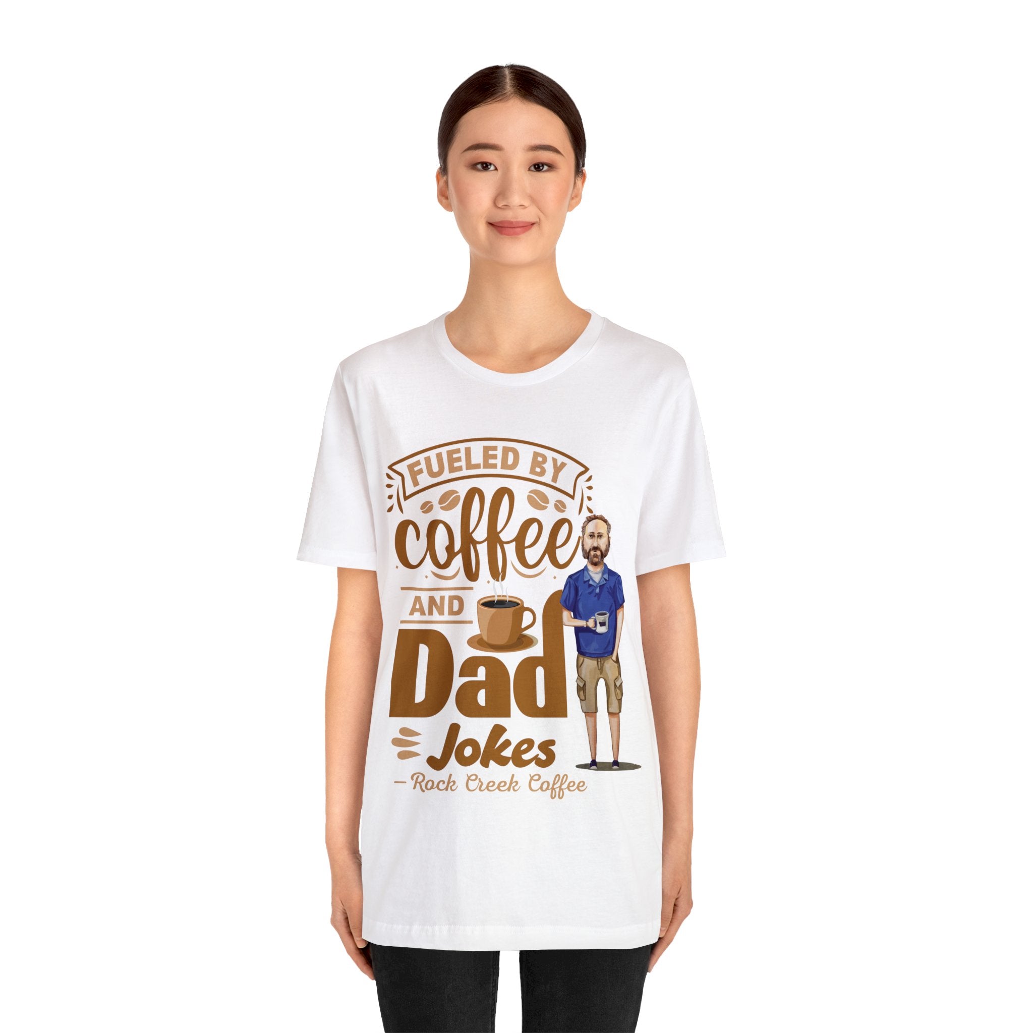 Woman wearing a white graphic T-shirt featuring the phrase “Fueled by Coffee and Dad Jokes” with a coffee cup illustration and cartoon dad character, standing against a plain white background.