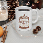 White Rock Creek Coffee mug featuring the phrase “Caffeine Because Adulting Is Hard” in brown and tan lettering, filled with coffee and styled with pinecones, cinnamon sticks, and soft holiday lights in the background.