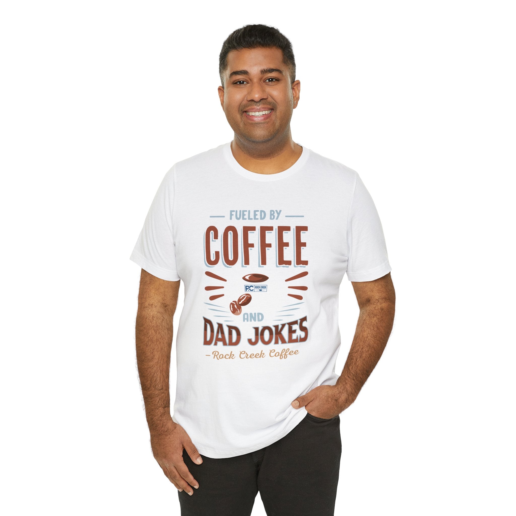 Adult man smiling while modeling a white short-sleeve T-shirt printed with a bold graphic that reads “Fueled by Coffee and Dad Jokes,” featuring a coffee cup illustration, shown against a plain white background.