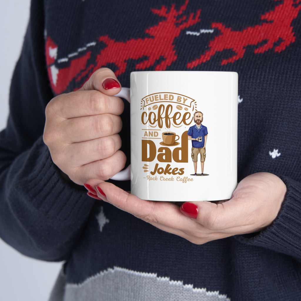 Person wearing a festive sweater holding a white ceramic mug that reads “Fueled by Coffee and Dad Jokes – Rock Creek Coffee,” featuring a cartoon illustration of a man holding a coffee cup.