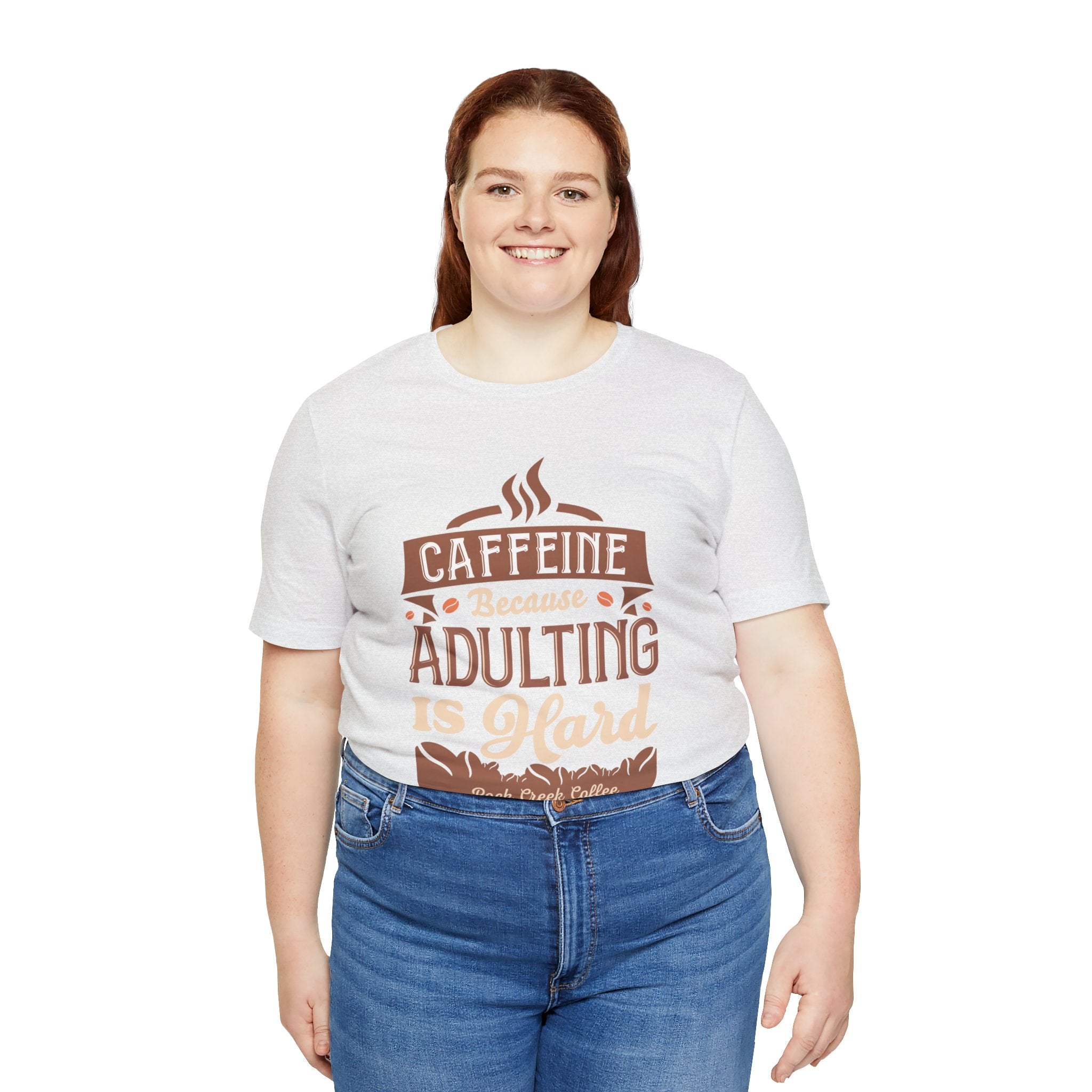 Adult woman smiling while modeling an ash short-sleeve T-shirt featuring the “Caffeine Because Adulting Is Hard” graphic in a stacked typographic design, shown against a plain white background.