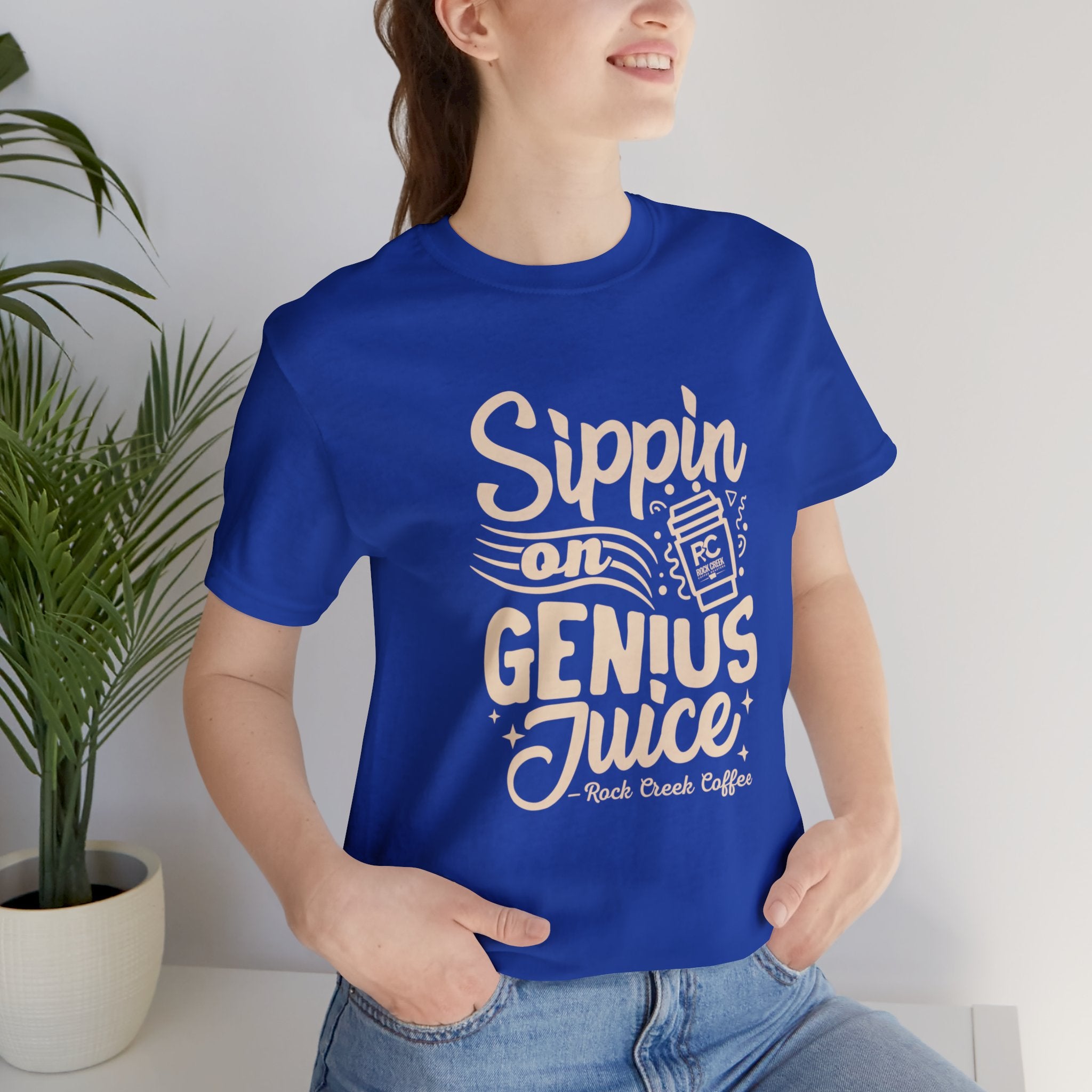 Smiling woman wearing a royal blue short-sleeve T-shirt printed with the slogan “Sippin on Genius Juice” and a stylized coffee to-go cup graphic, seated casually with indoor plants in the background.