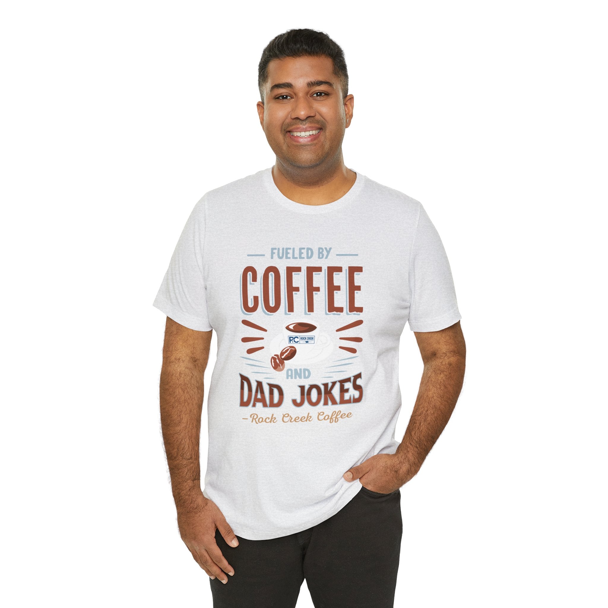 Adult man smiling while modeling an ash short-sleeve T-shirt featuring the “Fueled by Coffee and Dad Jokes” graphic with a coffee cup illustration, shown against a plain white background.