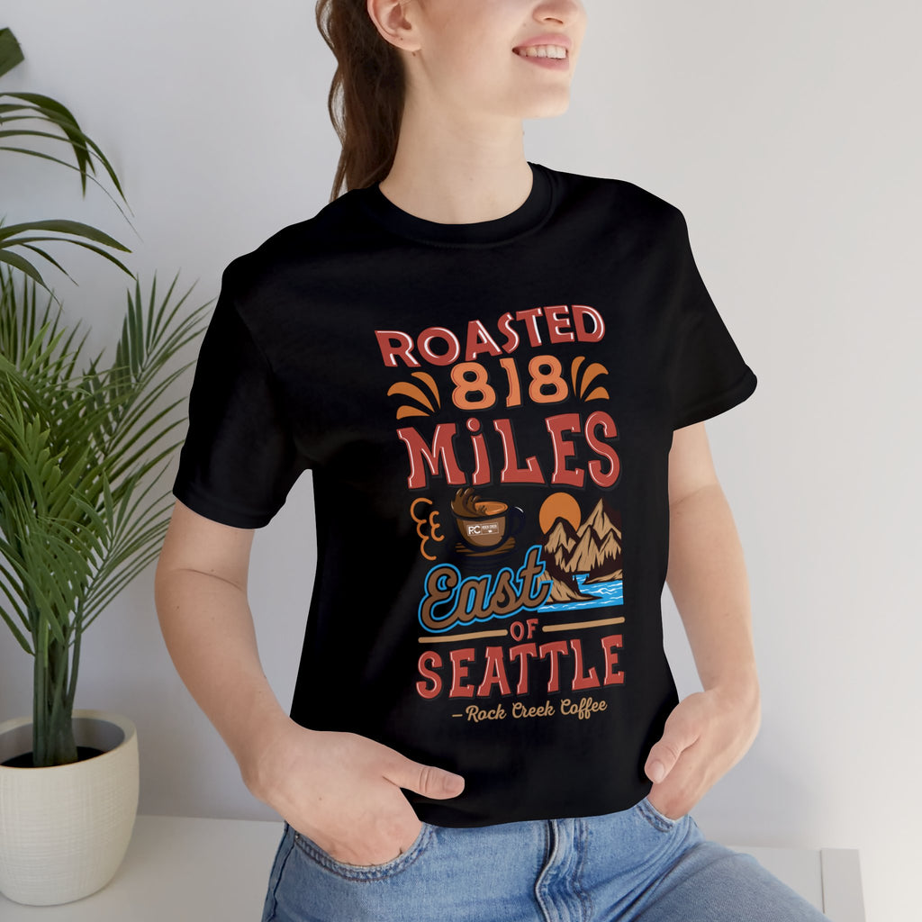 Adult woman modeling a black short-sleeve T-shirt printed with a colorful graphic that reads “Roasted 818 Miles East of Seattle,” featuring a steaming coffee cup and mountain lake illustration, shown in an indoor setting with a potted plant and light background.