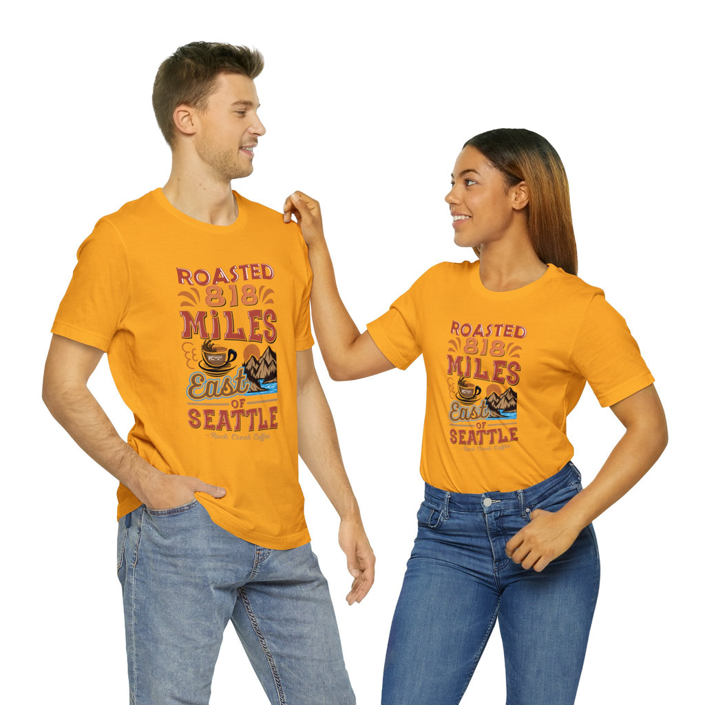 Adult man and woman modeling matching golden yellow short-sleeve T-shirts printed with a colorful graphic that reads “Roasted 818 Miles East of Seattle,” featuring a steaming coffee cup and mountain lake illustration, shown against a plain white background.