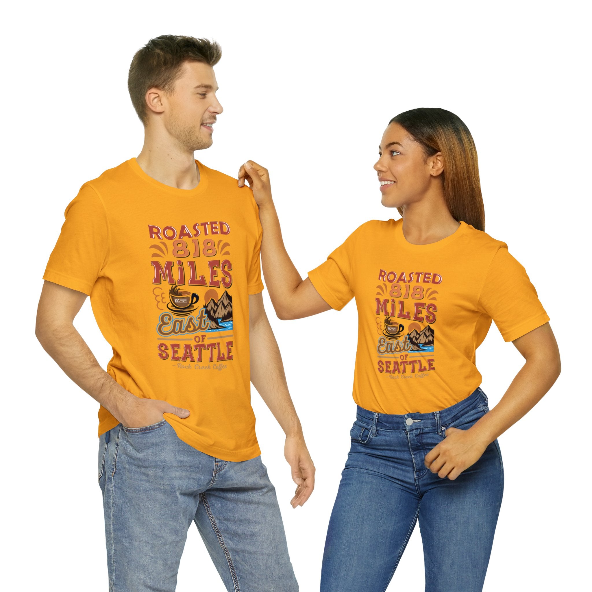 Adult man and woman modeling matching golden yellow short-sleeve T-shirts printed with a colorful graphic that reads “Roasted 818 Miles East of Seattle,” featuring a steaming coffee cup and mountain lake illustration, shown against a plain white background.