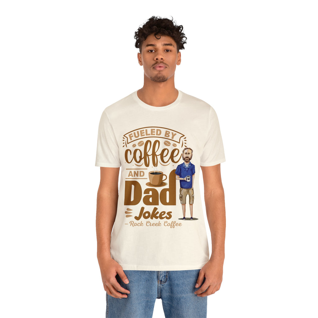 Young man modeling a cream short-sleeve T-shirt with a coffee-themed printed graphic reading “Fueled by Coffee and Dad Jokes,” showing a steaming coffee cup and a cartoon dad holding a mug, on a plain white background.