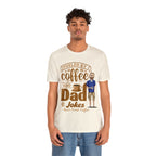 Young man modeling a cream short-sleeve T-shirt with a coffee-themed printed graphic reading “Fueled by Coffee and Dad Jokes,” showing a steaming coffee cup and a cartoon dad holding a mug, on a plain white background.