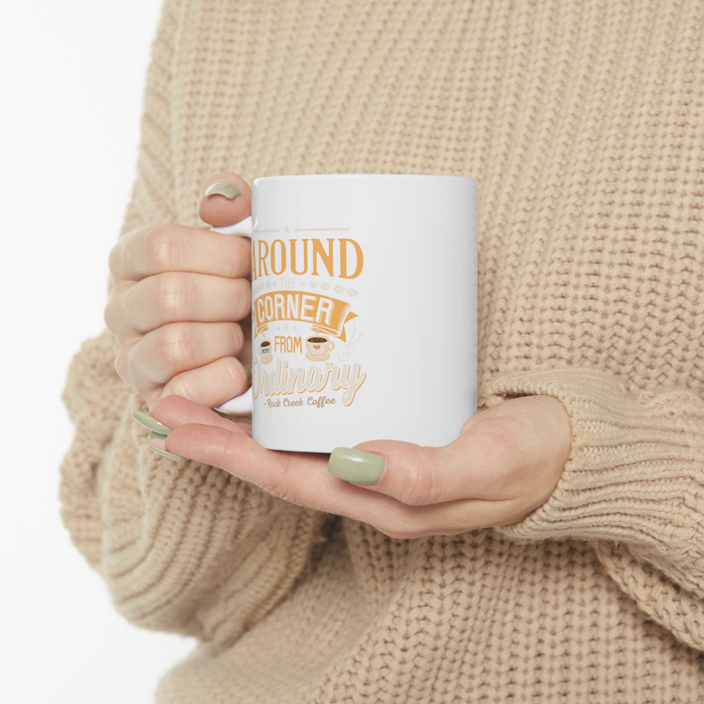 Person wearing a beige knit sweater holding a white Rock Creek Coffee mug with “Around the Corner from Ordinary” design in gold and cream lettering.