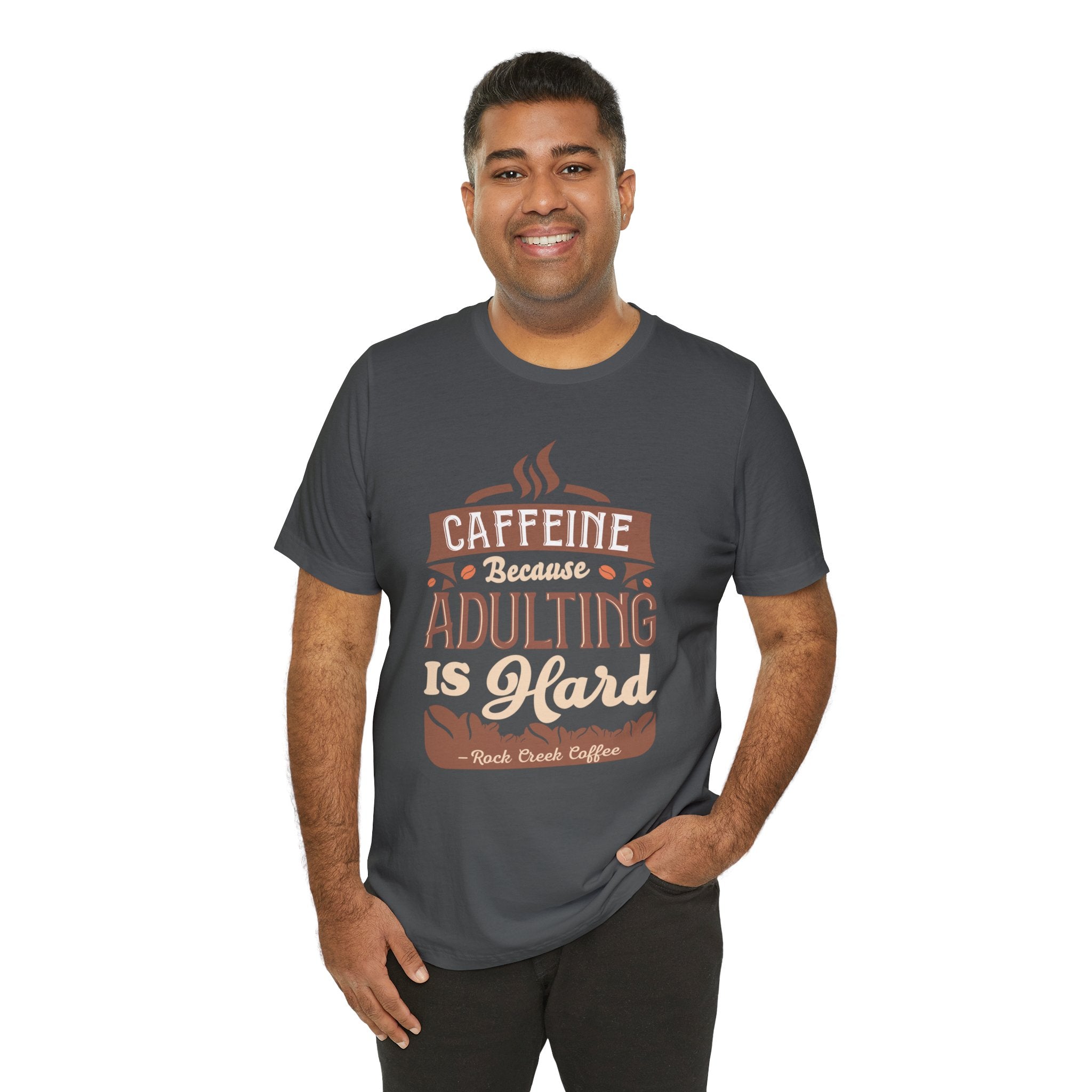 Adult man smiling while modeling an asphalt short-sleeve T-shirt featuring the “Caffeine Because Adulting Is Hard” graphic in a stacked typographic design, shown against a plain white background.