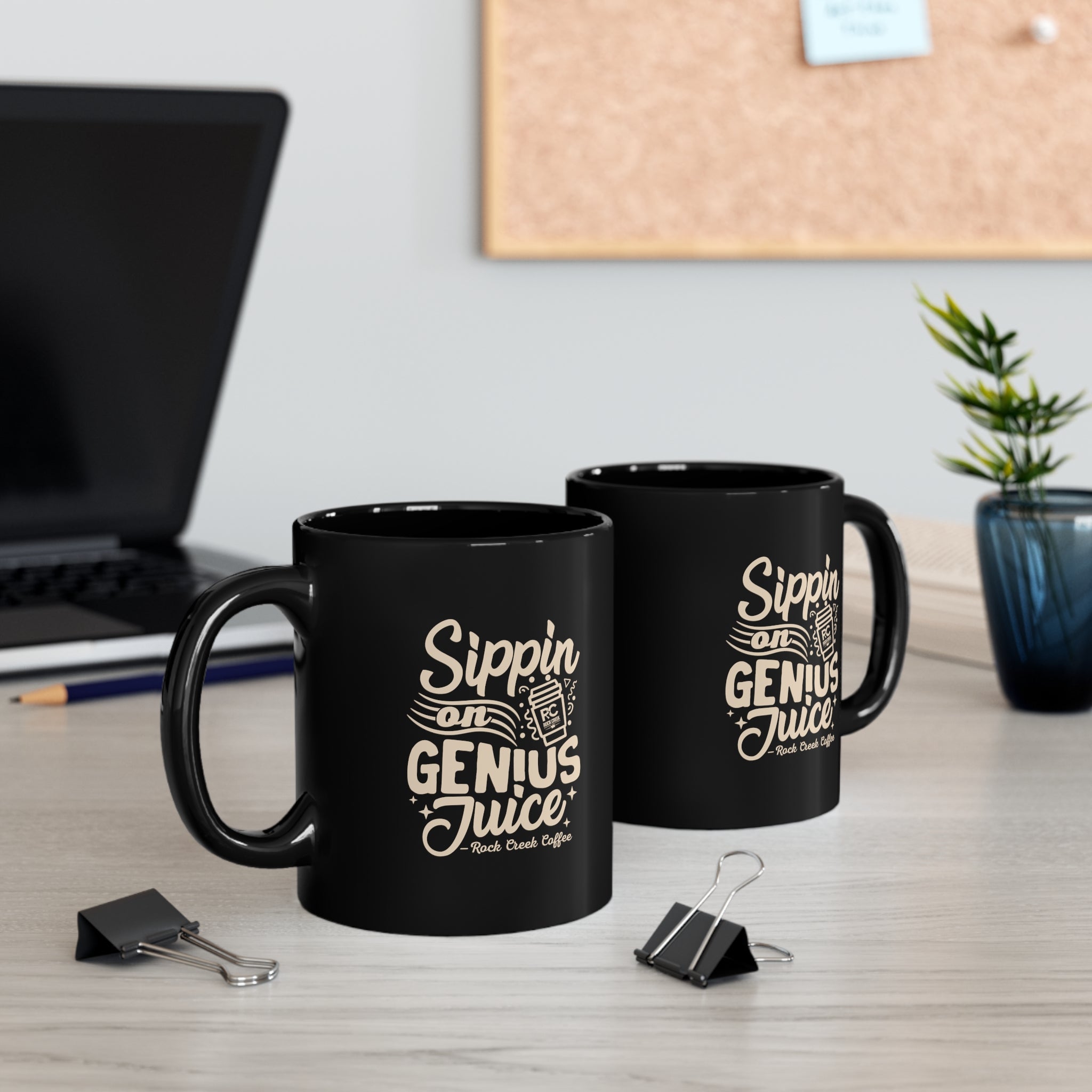 Two black Rock Creek Coffee mugs with “Sippin on Genius Juice” design in cream lettering displayed on a desk with a laptop, binder clips, stacked books, and a small potted plant in the background.